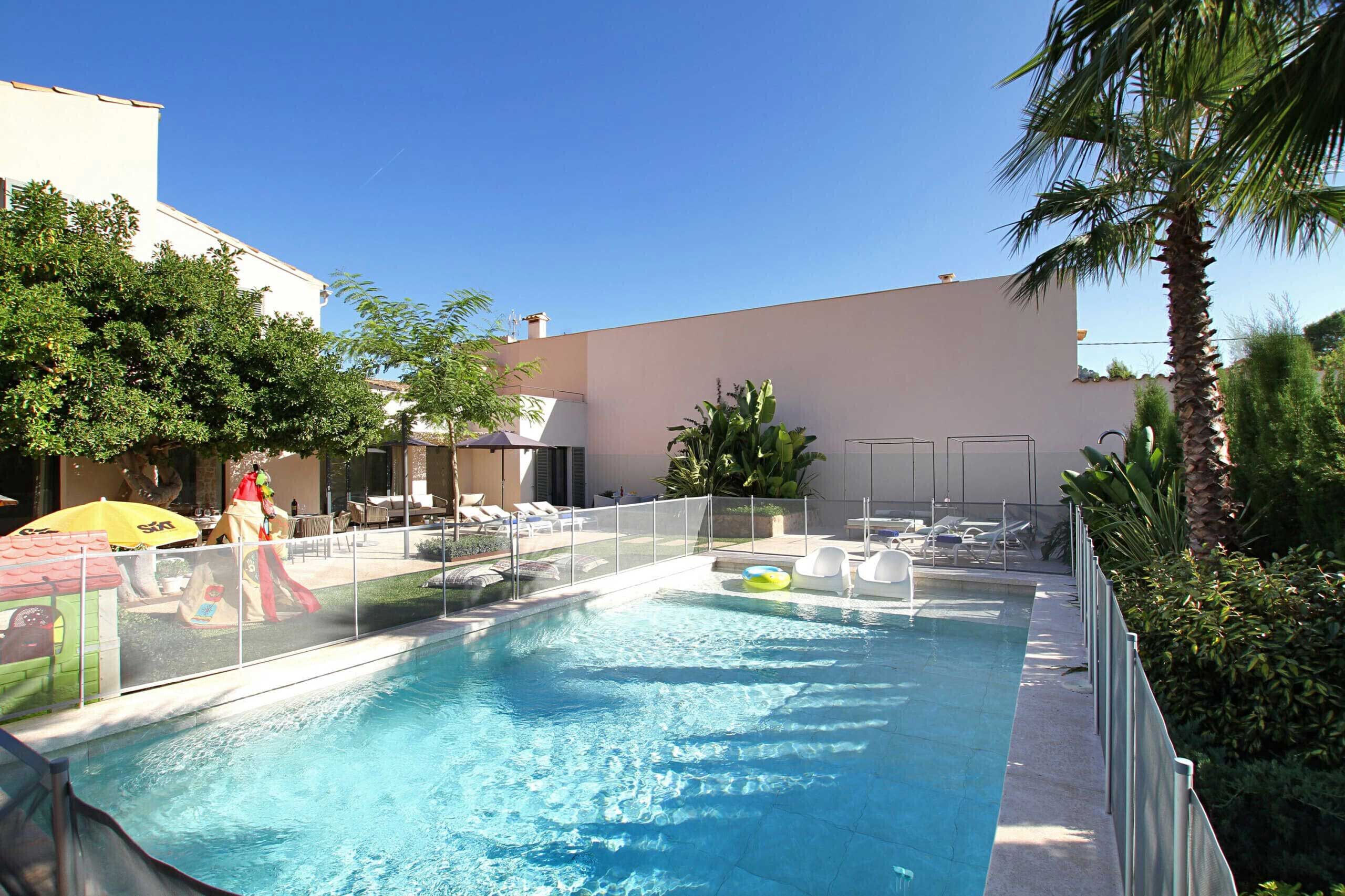 A swimming pool with lounge chairs and umbrellas, surrounded by greenery and a building.