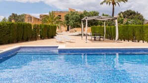 Ein Swimmingpool mit gefliestem Deck, einem überdachten Sitzbereich und Blick auf ein traditionelles Gebäude im Hintergrund.