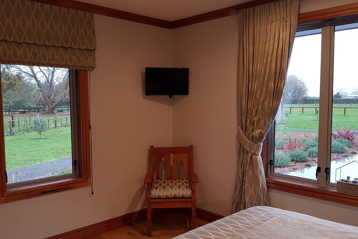 A bedroom with a bed, a chair, a TV mounted on the wall, and large windows with curtains.