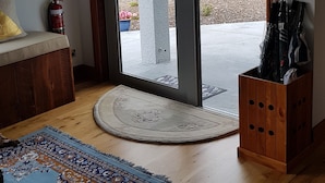 A room with a wooden floor, a patterned rug, a bench, a fire extinguisher, and a view of a garden through a glass door. - BRUNTWOODLODGE - Adults Only (Tamahere)