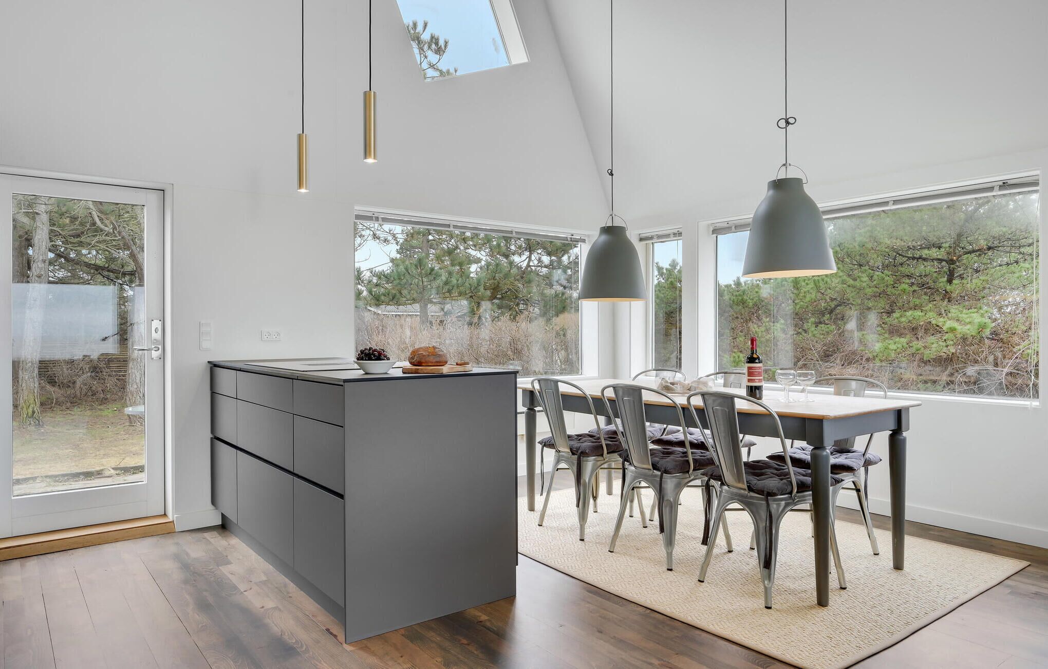 Een moderne keuken met een centraal kookeiland, grote ramen en een eethoek met stoelen en een tafel.
