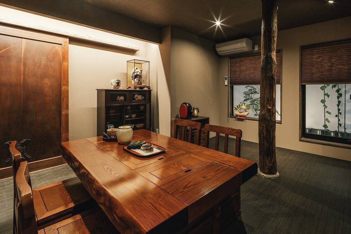 Uma sala de jantar com mesa de madeira, cadeiras, um armário com objetos decorativos e uma janela com uma planta.