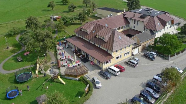 Ein großes Gebäude mit mehreren Dächern, umgeben von grünen Feldern und Bäumen. Es gibt einen Spielplatz mit Rutsche und Trampolin. Vor dem Gebäude sind mehrere Autos geparkt.