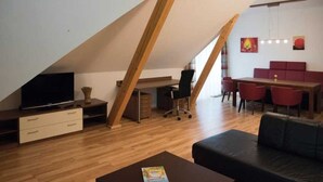 Ein Loft-Wohnraum mit schrägem Dach, Holzbalken, einem Fernseher, einem Schreibtisch, einem schwarzen Bürostuhl, einem roten Essplatz und einer schwarzen Ledersofa.