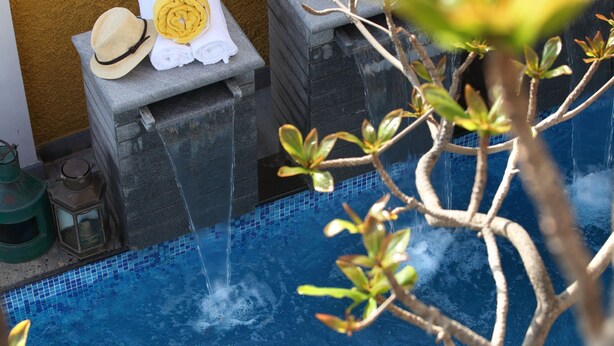 A pool area with a water feature, a towel and hat on a ledge, and a plant in the foreground. - Waterfront Granary A Museum Hotel (Kochi)