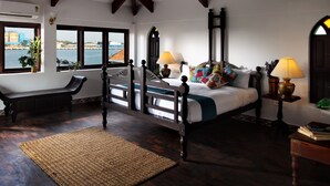 A bedroom with a large bed, two bedside tables with lamps, a window with a view, a ceiling fan, and a wooden floor. - Waterfront Granary A Museum Hotel (Kochi)