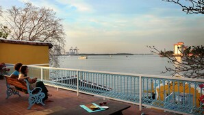 A balcony with a view of a river, a bench, and a table with a plate and drinks. - Waterfront Granary A Museum Hotel (Kochi)