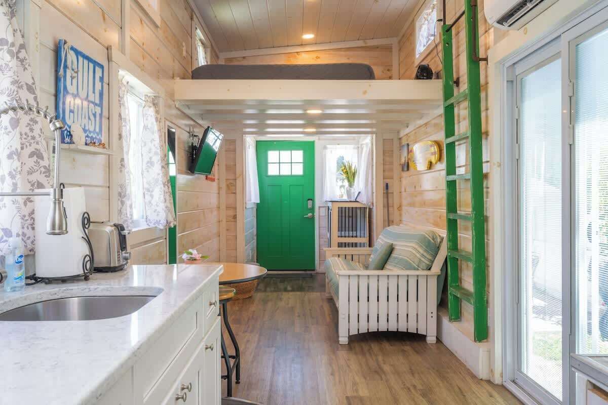 A compact kitchen with a sink, a toaster, and a green door leading to a small seating area with a bench and a ladder.