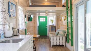 A compact kitchen with a sink, a toaster, and a green door leading to a small seating area with a bench and a ladder.