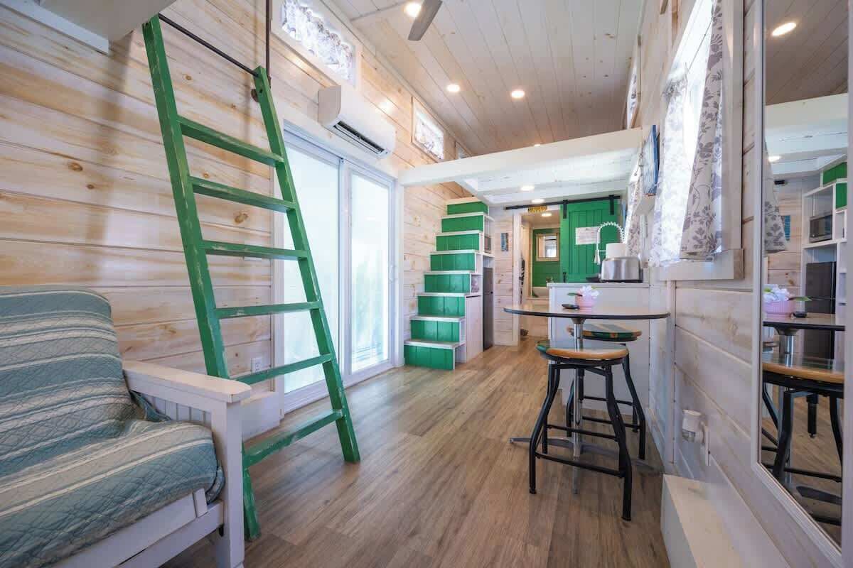 A cozy cabin interior with a wooden floor, a green ladder, a striped sofa, and a kitchen area with green cabinets.
