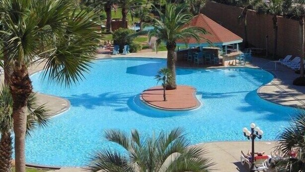 A resort pool with palm trees, lounge chairs, and a view of the ocean. - A Wave From It  All! (Galveston)
