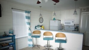 A kitchen with white cabinets, a microwave, a refrigerator, and three wicker bar stools. - A Wave From It  All! (Galveston)