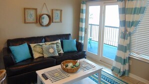 A living room with a dark brown sofa, a white coffee table with a basket, and a view of a balcony with striped curtains. - A Wave From It  All! (Galveston)