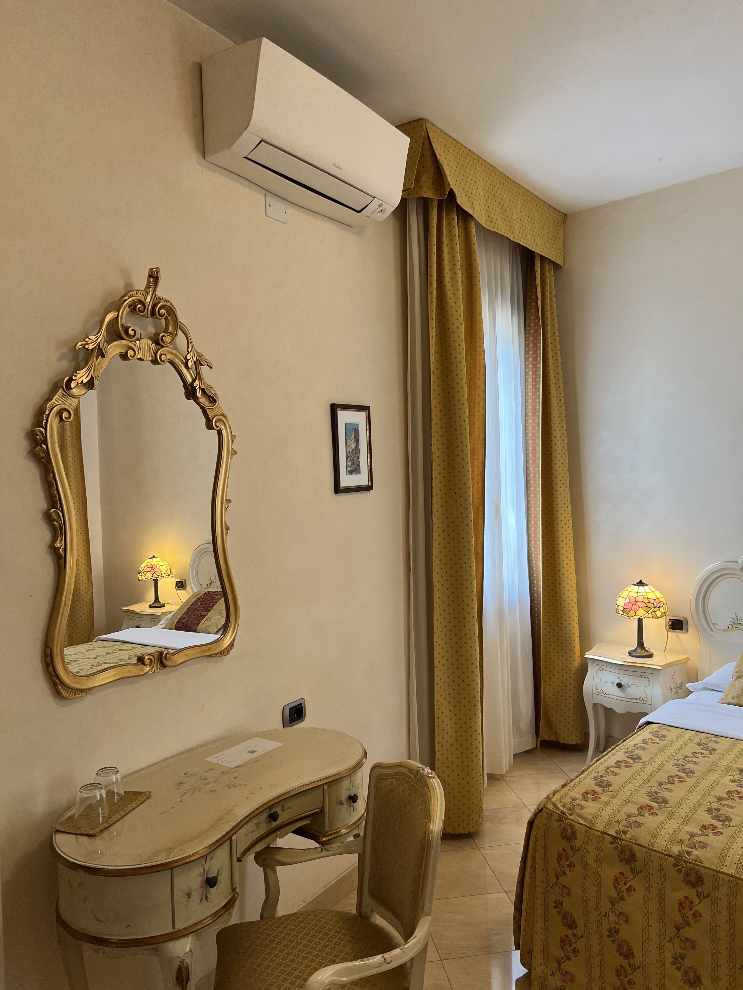 A hotel room with a bed, a desk, a chair, an ornate mirror, an air conditioner, and a nightstand with a lamp.