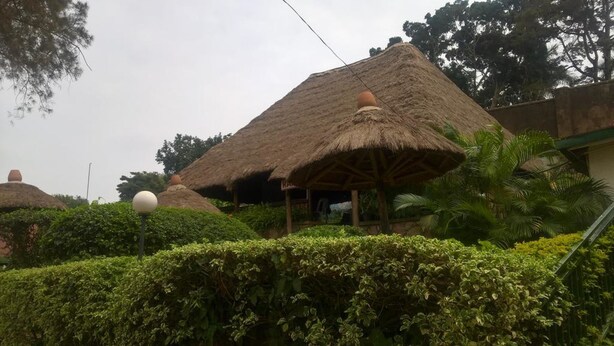 Een traditionele constructie met rieten dak en een overdekte veranda, omgeven door goed onderhouden hagen en palmbomen.