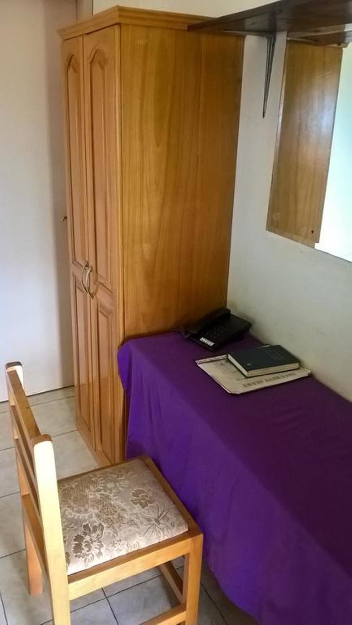 Una habitación pequeña con un armario de madera, una cama morada, una silla de madera y una mesita con libros y un teléfono.