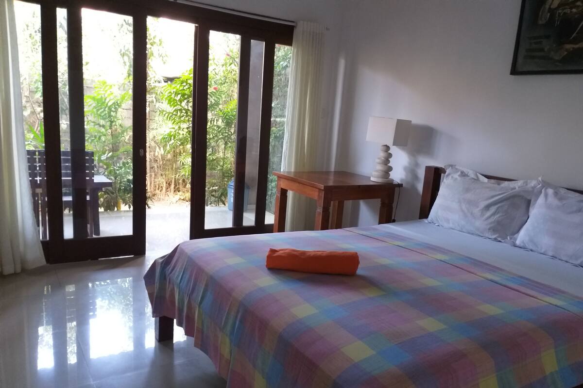 A room with a bed, a table, a lamp, and a view of greenery outside.