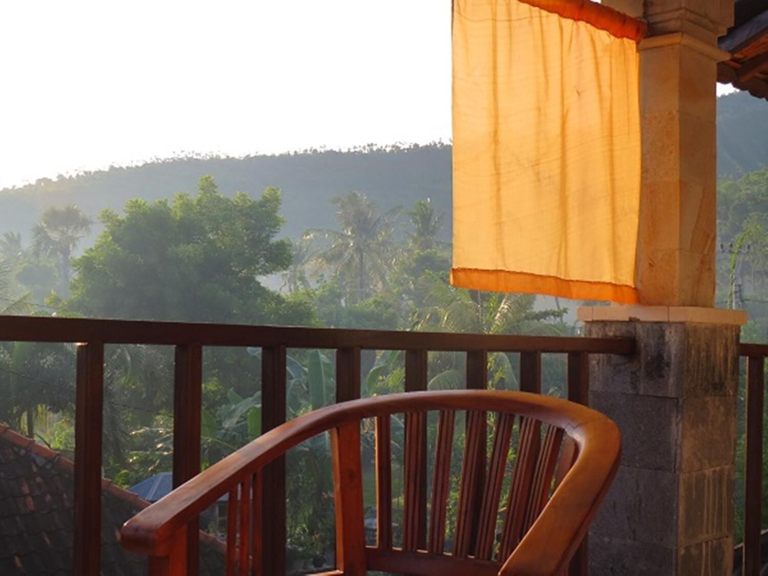 A wooden chair on a balcony with a view of a hilly landscape and palm trees.