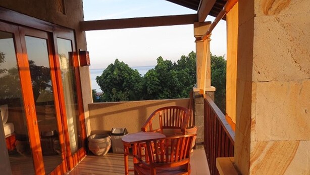 A balcony with a wooden table and chairs, a view of greenery, and a stone wall. - Bali Sari Homestay (Karangasem)