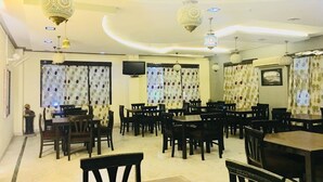 A dining area with multiple tables and chairs, patterned curtains, and hanging light fixtures. - Hotel Chanakya (Agra)