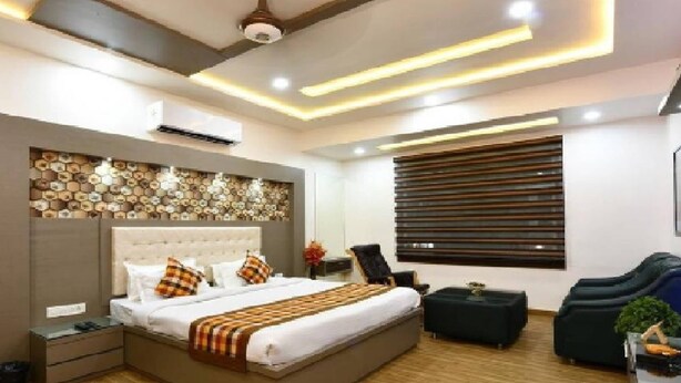 A modern bedroom with a large bed, a headboard with a decorative wall, a window with blinds, a dark sofa, and a ceiling fan. - Hotel Chanakya (Agra)
