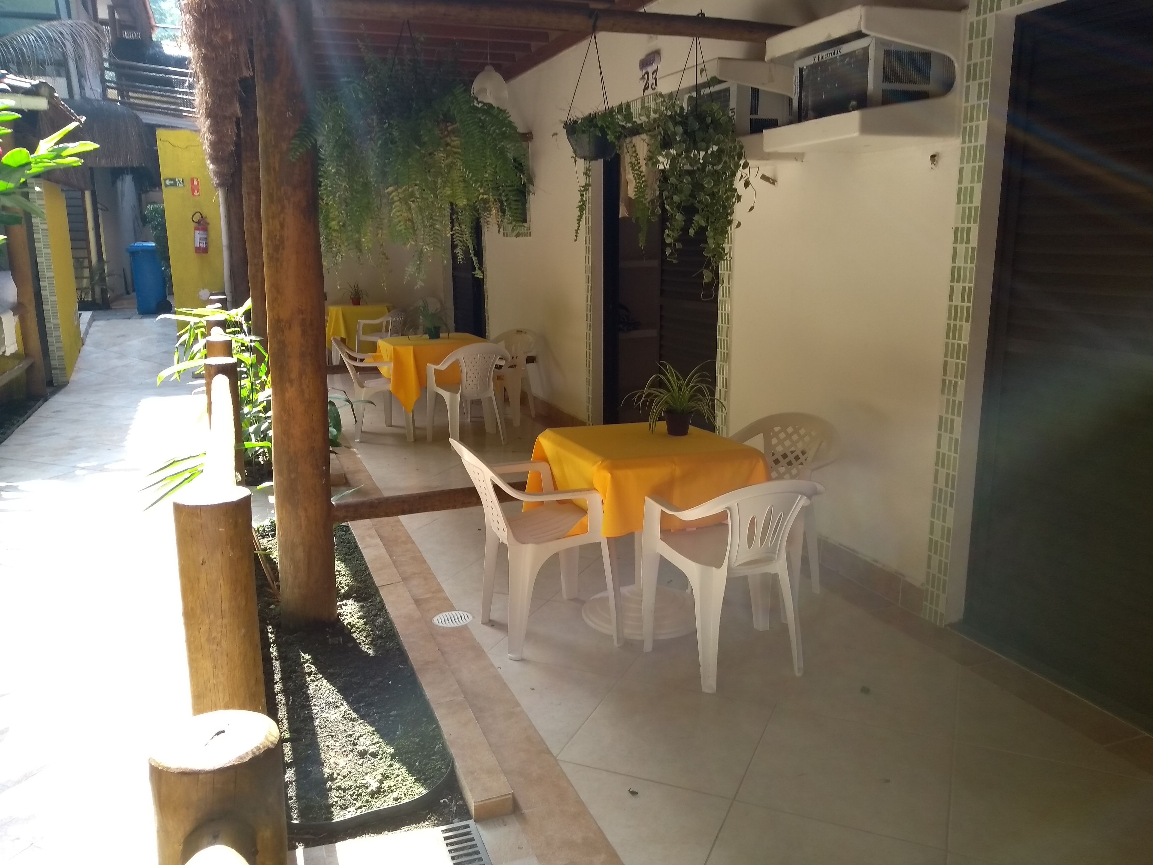 Área de refeições ao ar livre com mesas e cadeiras, rodeada por plantas e um chão de azulejos.