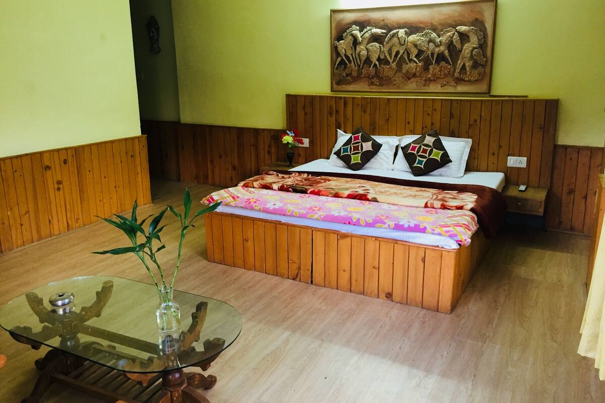 A wooden bed with a colorful bedspread, a glass table with a vase, and a painting on the wall.