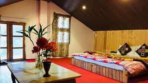 Een knus interieur met een houten plafond, een groot raam met bloemen gordijnen, een houten bed met kleurrijk beddengoed, en een tafel met een bloevas.