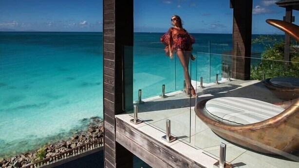 A glass balustrade overlooking a turquoise sea, a modern outdoor seating area, and a clear sky.
