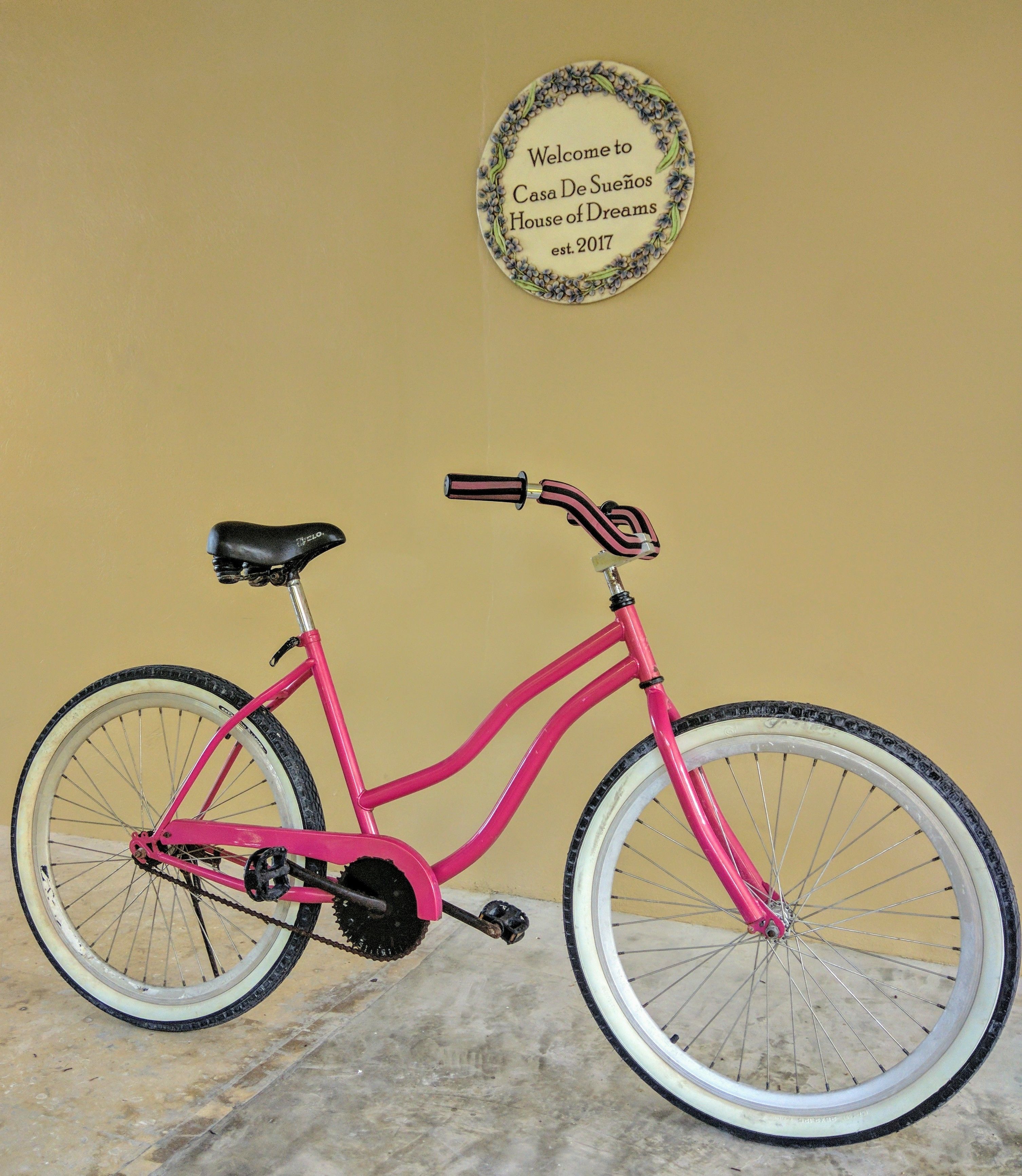A pink bicycle parked against a yellow wall with a welcome sign above it.