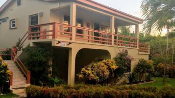 Two-story building with a flat roof and balcony, featuring windows and a staircase with a red railing, set amidst a well-maintained garden.
