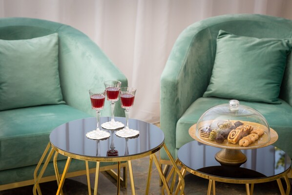 A room with two green sofas, a round black table with gold legs, and a round table with a glass dome displaying pastries. - Hotel Corner (Split)