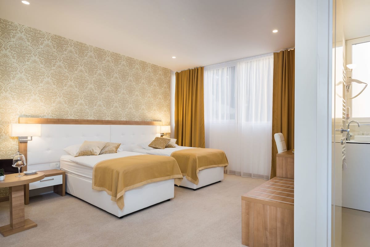 A hotel room with two beds, a nightstand, a desk, a chair, a small table, a window with curtains, and a bathroom with a sink and mirror.