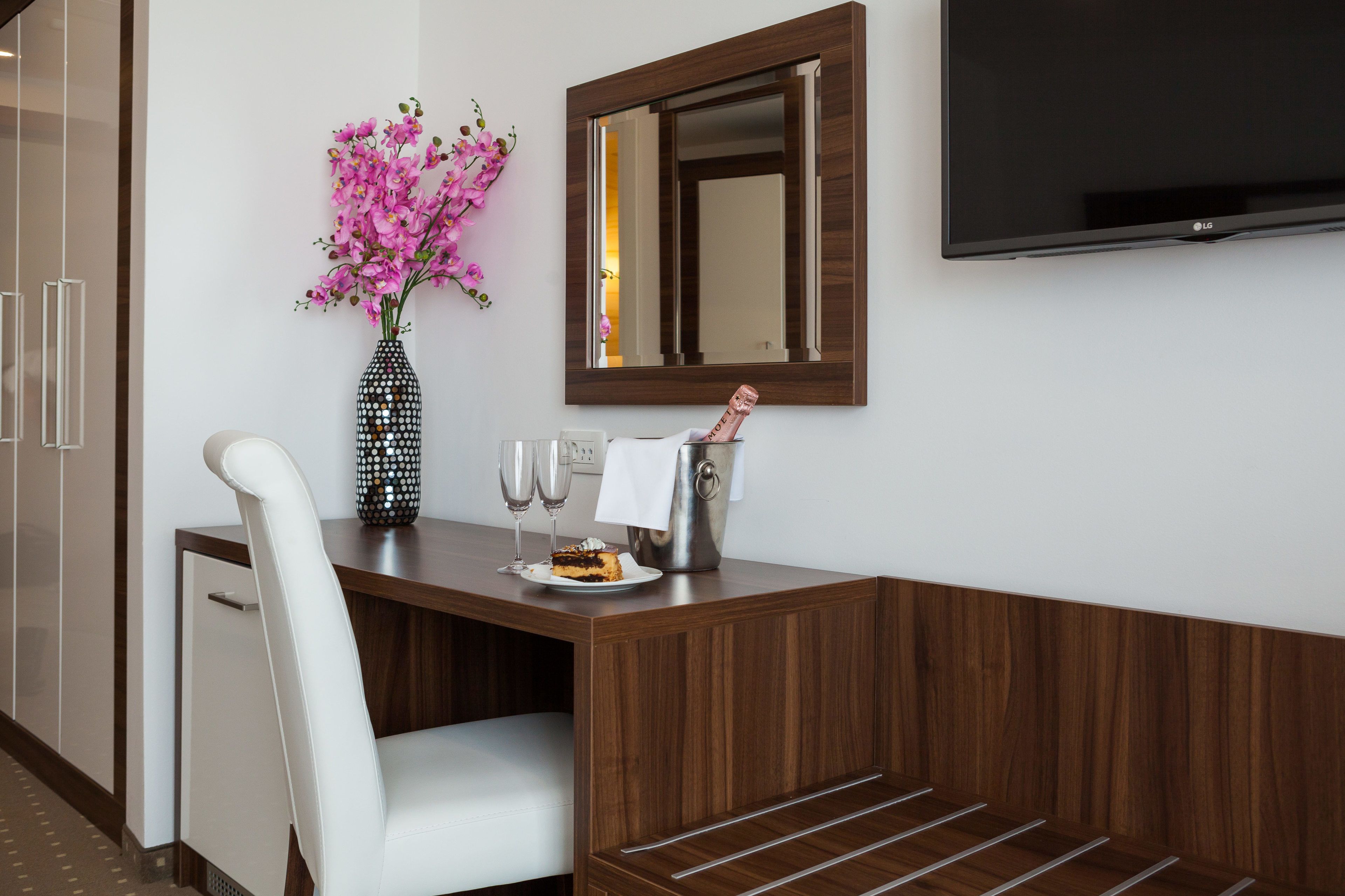 Ein modernes Hotelzimmer mit einem Holztisch, einem weißen Stuhl, einer Vase mit rosa Blumen, einer Champagnerflasche und einem Flachbildfernseher an der Wand.