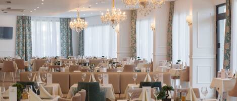 A spacious dining area with multiple tables and chairs, elegant chandeliers, and large windows with curtains.