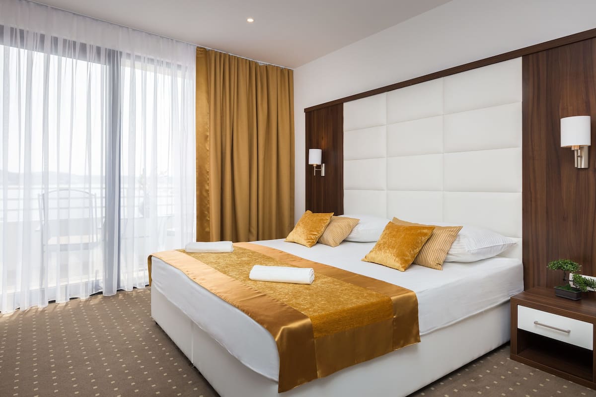 A hotel room with a large bed, a bedside table with a lamp, and a view of the outdoors through sheer curtains.