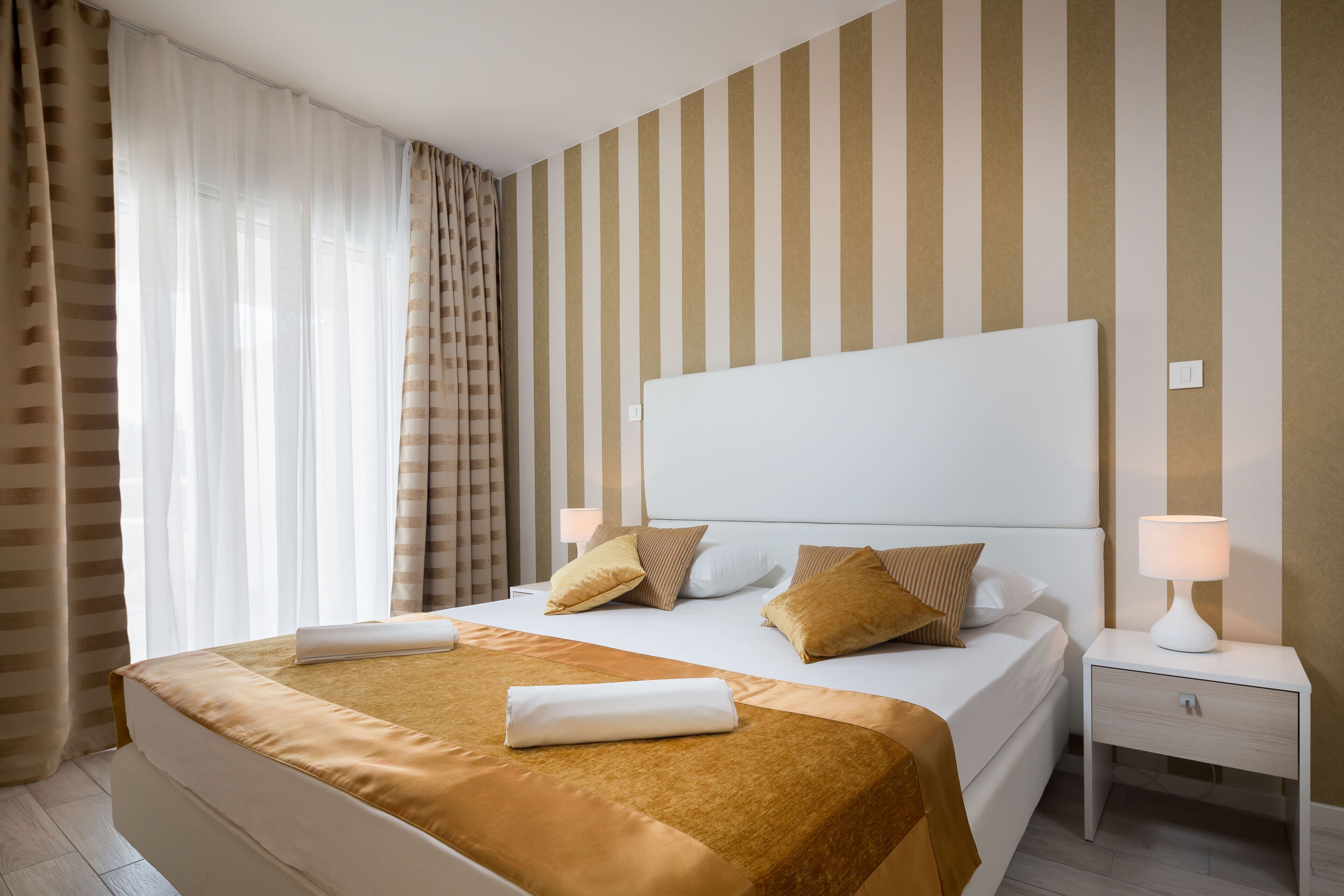 A neatly made bed with a white headboard, a bedside table with a lamp, and striped wallpaper.