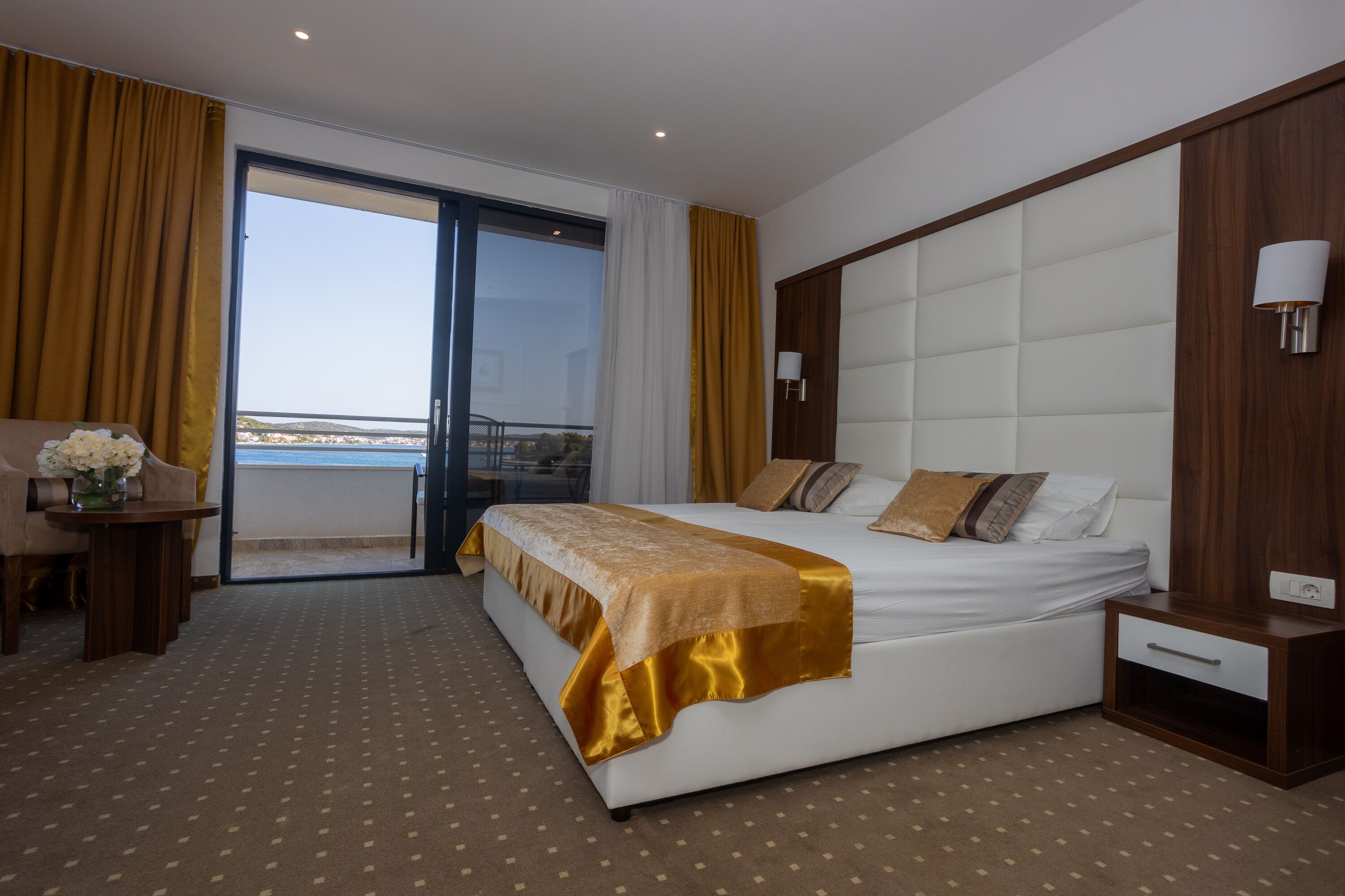 A hotel room with a large bed, a bedside table, a nightstand, a lamp, and a view of the outdoors.
