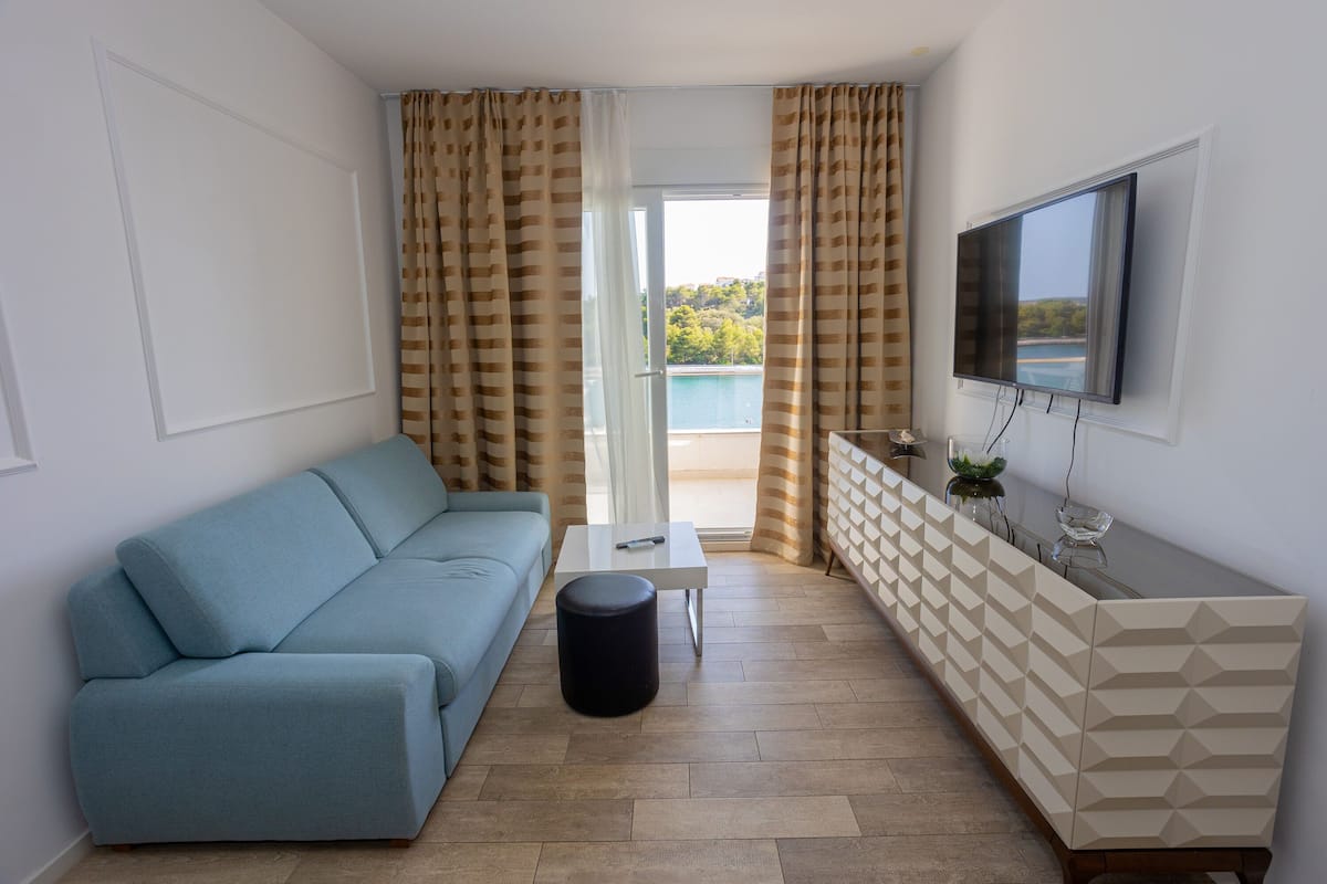 A modern living room with a sofa, a small table, a TV mounted on the wall, and a view of a pool and greenery outside.