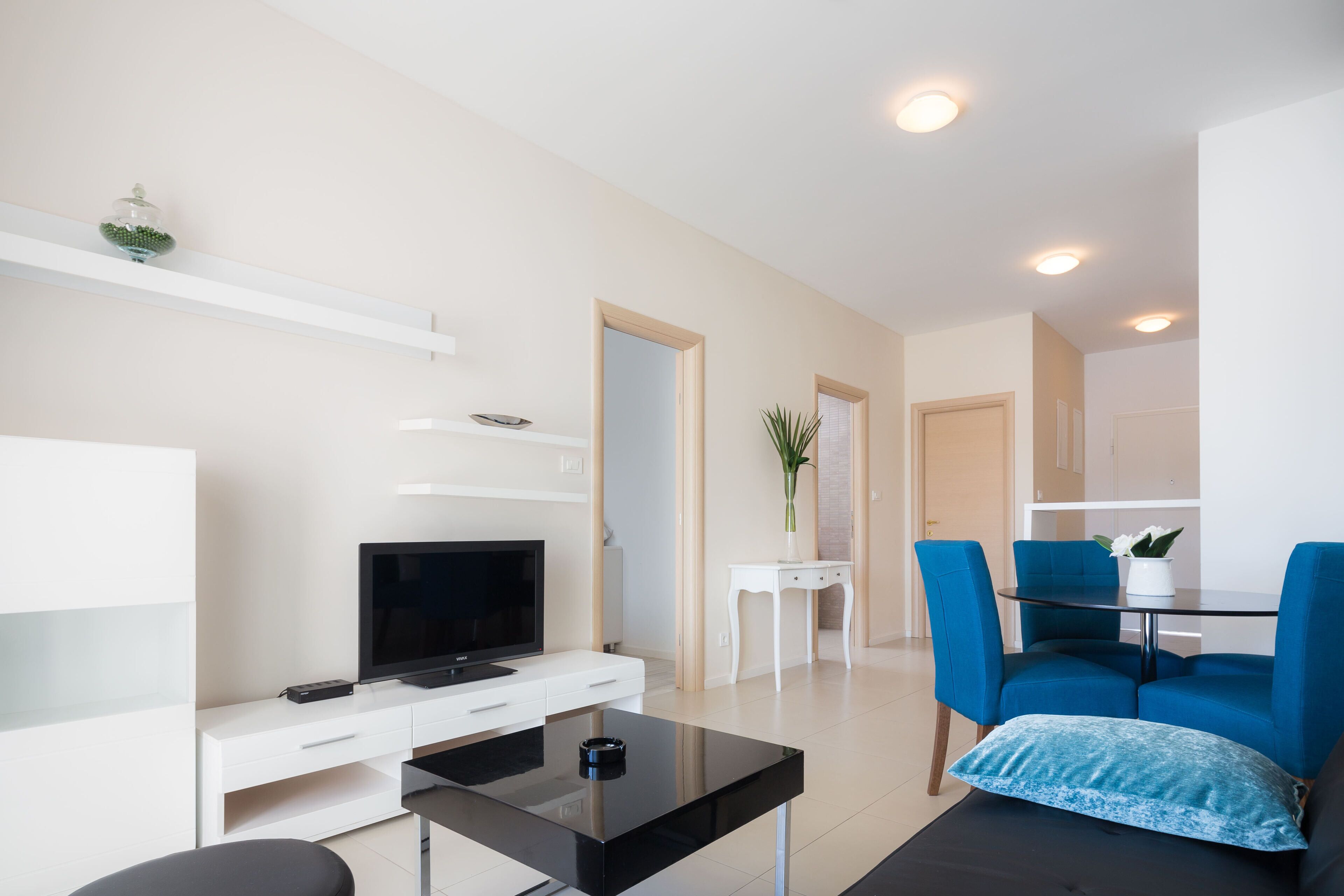 Een moderne woonkamer met een flatscreen televisie, een salontafel, een bank en een eethoek met stoelen.