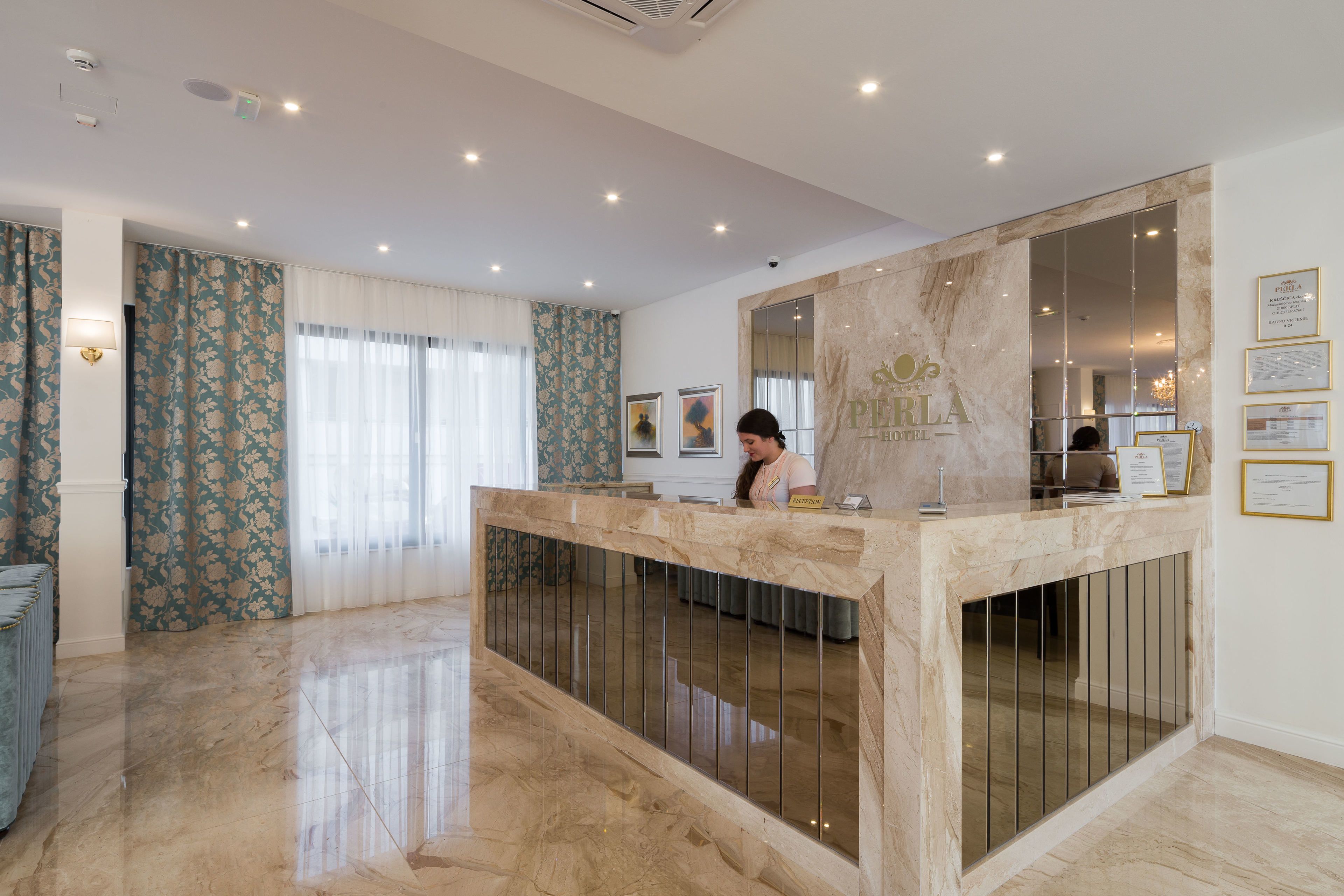 Zona de recepción de hotel con mostrador de mármol, certificados enmarcados en la pared y un amplio ventanal con cortinas.