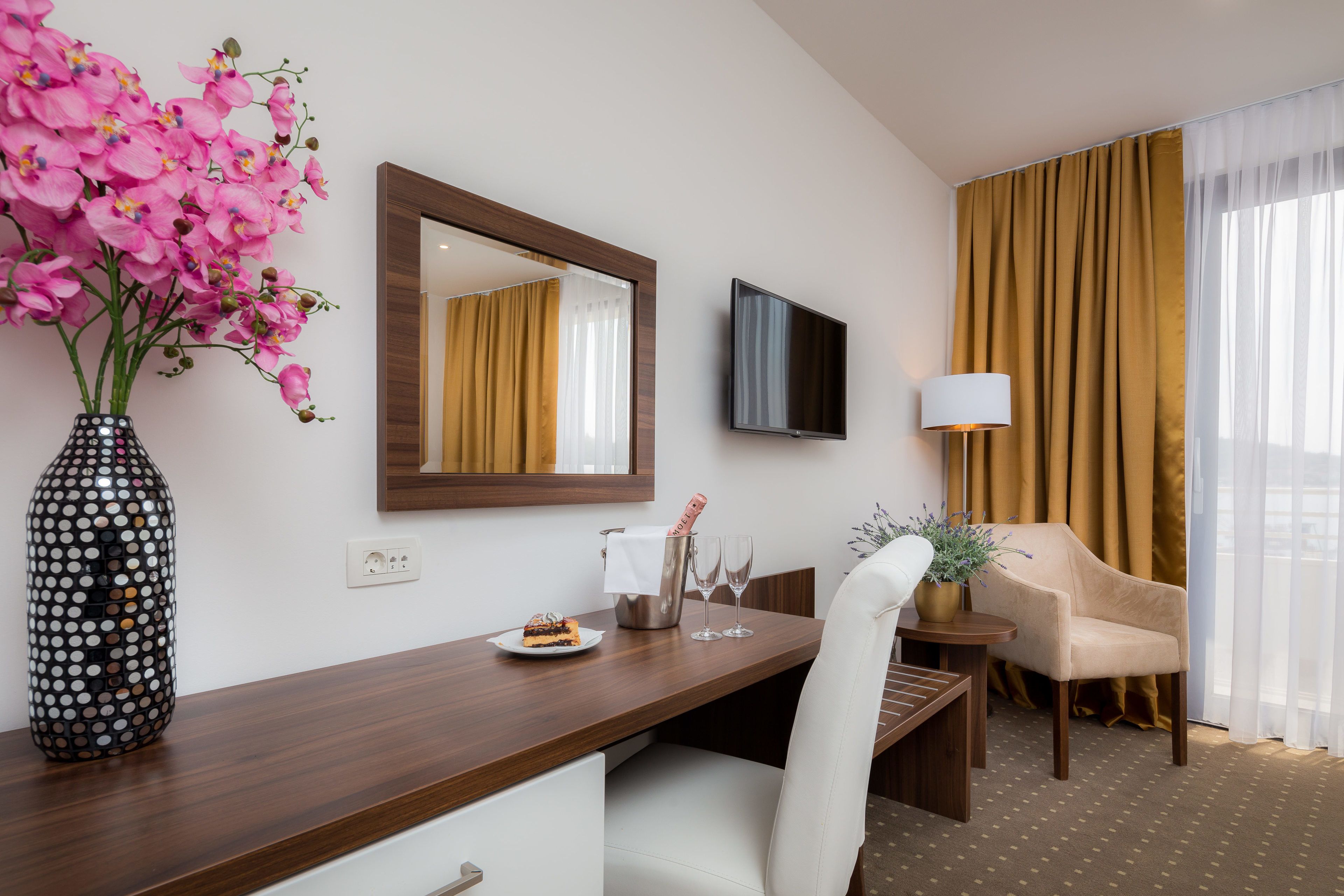 Une chambre d’hôtel avec un bureau en bois, un vase rempli de fleurs roses, un téléviseur à écran plat, une lampe, une chaise et une grande fenêtre avec des rideaux.