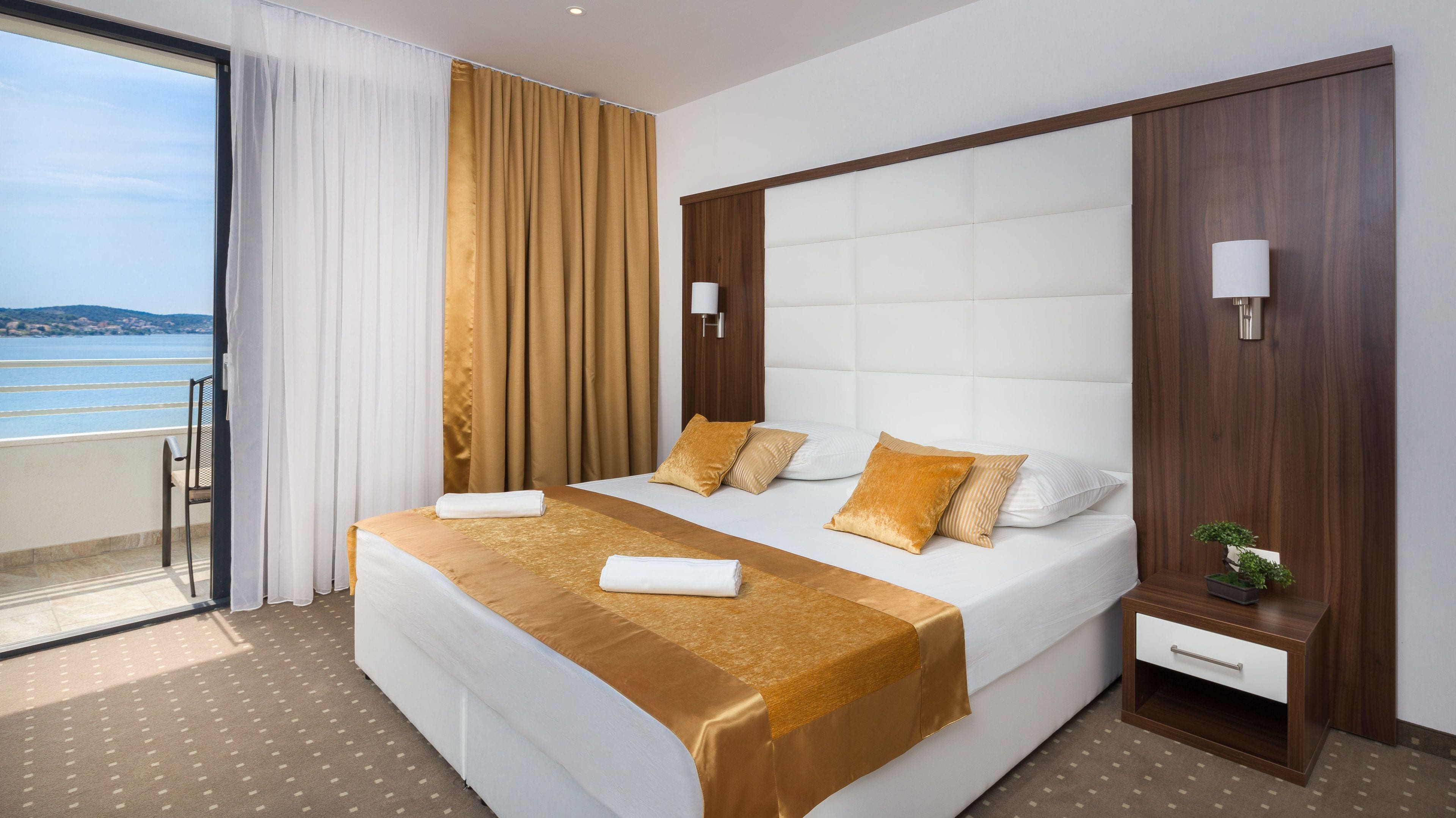 A hotel room with a large bed, a small bedside table, a nightstand, a view of the sea, and a balcony.