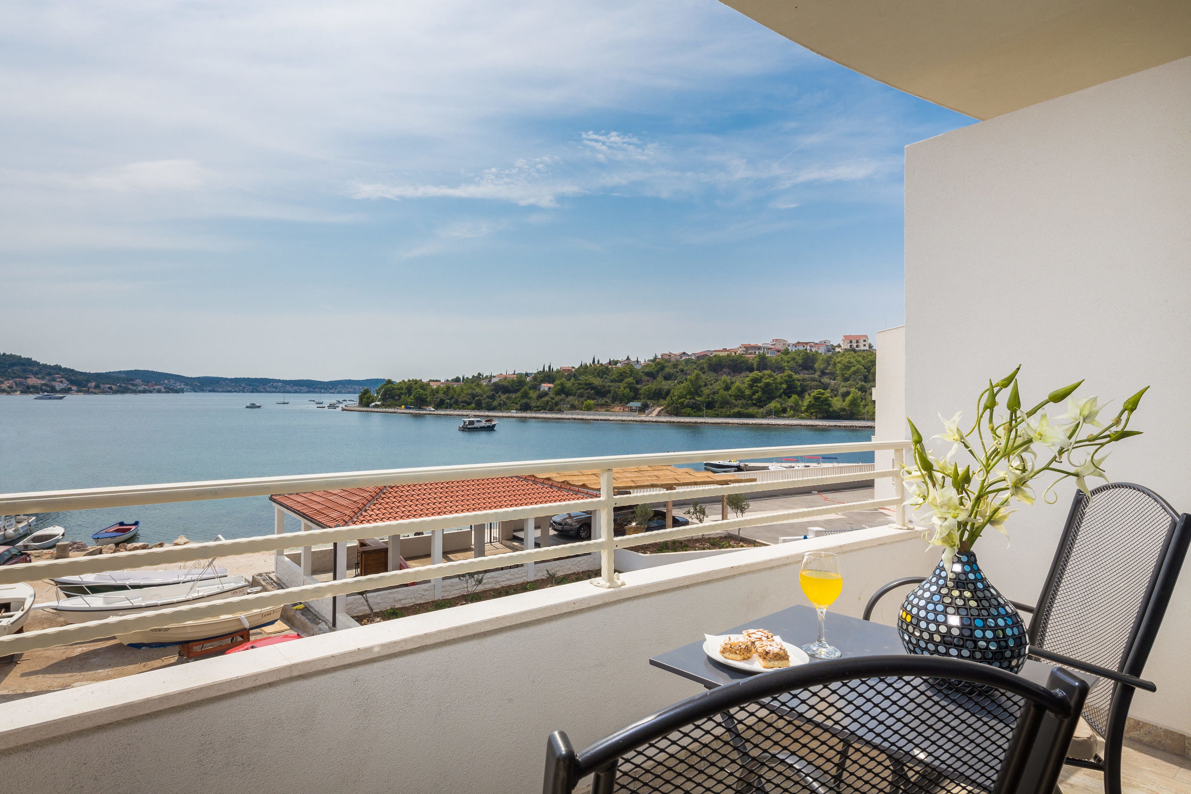 Een balkon met een tafel, een stoel, een vaas met bloemen, een glas drinken en uitzicht op zee met boten en gebouwen in de verte.