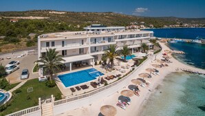 Un hotel de varios pisos con piscina, sillas de descanso en la playa, sombrillas y vistas al mar.