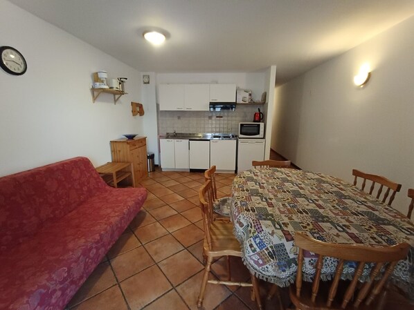 A compact living space with a red sofa, a dining table with chairs, a kitchen area with white cabinets, and a clock on the wall.