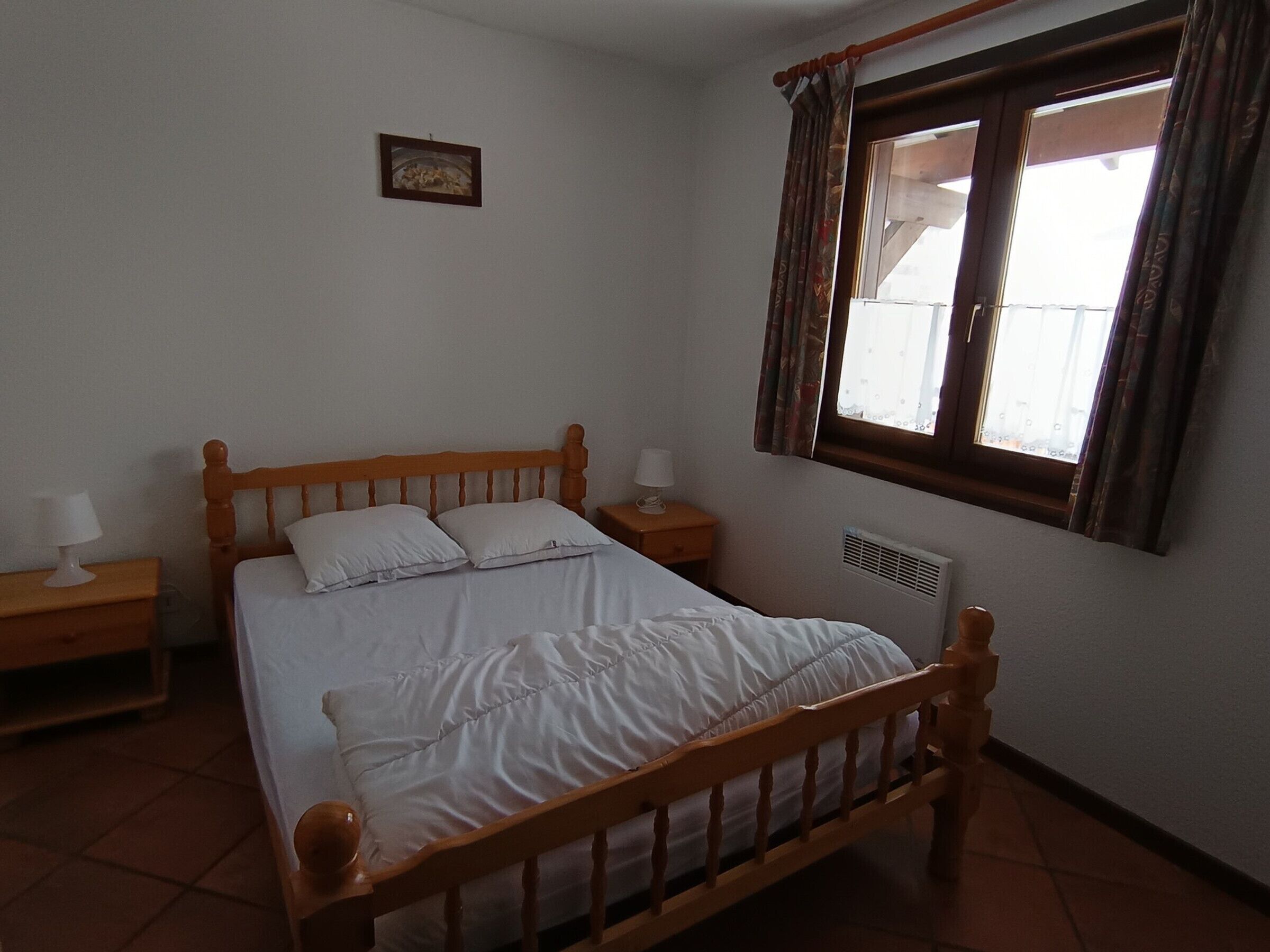 A bedroom with a wooden bed, a nightstand with a lamp, a window with curtains, and a radiator.