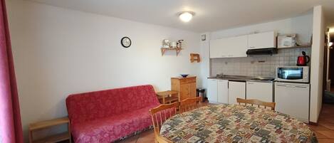 A compact living space with a kitchenette, a red sofa, a dining table with chairs, and a patterned tablecloth.