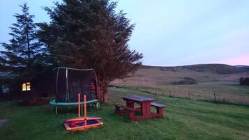 A grassy outdoor area with a trampoline, picnic table, and a covered play structure.