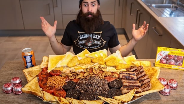 Eine Person sitzt an einem Tisch mit einer großen Auswahl an Speisen, darunter Toast, Würstchen, Bohnen und Schokolade.